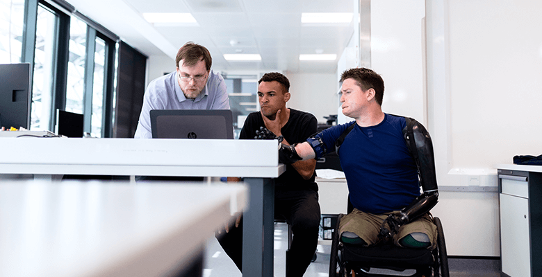 3 men, including a man in a wheelchair with prosthetics, look at a laptop.